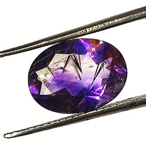 Peut inclure: Une pierre précieuse taille ovale présentant un dégradé de tons violet profond à transparent avec des inclusions sombres en forme d'aiguilles visibles sur toute la surface facettée. La pierre est tenue par des pinces métalliques.