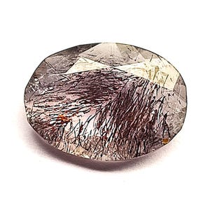 May include: An oval-shaped, faceted gemstone with a translucent, clear base. The interior is filled with dense, dark brown to black needle-like inclusions that create a textured, fibrous appearance throughout the stone's crystalline structure.