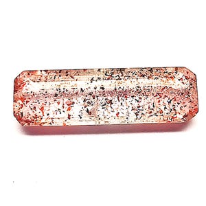 Puede incluir: Una gema rectangular con un tono rosa rojizo e inclusiones internas de partículas negras y rojas. La gema tiene un corte facetado con una superficie pulida, sobre un fondo blanco.