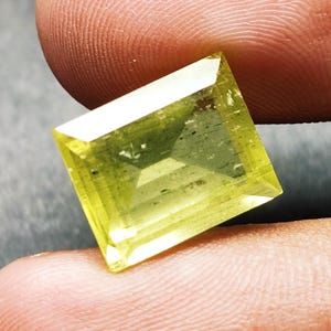 Könnte beinhalten: Ein rechteckiger facettierter Edelstein in leuchtendem Limettengrün mit einer transparenten, polierten Oberfläche. Der Stein weist scharfe, klare Kanten und einen geometrischen Schliff auf, der das Licht reflektiert und innere Klarheit zeigt.