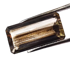 May include: A rectangular, faceted gemstone with a gradient of brown and golden-yellow tones held by metal tweezers. The stone features a clear, elongated shape with visible internal striations and a polished surface that reflects light.