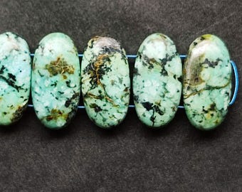 May include: A strand of oval-shaped turquoise beads with black and brown veining. The beads are strung on a blue cord, and the background is a dark gray fabric. These beads are suitable for jewelry making.