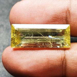 Può includere: Una gemma gialla rettangolare con taglio baguette e superficie trasparente sfaccettata. La pietra presenta una vivace tonalità giallo limone con inclusioni interne visibili e una forma geometrica netta e definita.
