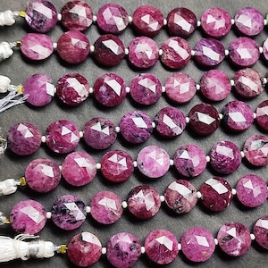 May include: Rows of faceted, round, ruby-colored gemstone beads. The beads are strung on white string, with white tassels visible at the ends. The gemstones have a mix of deep red and pink hues, with reflective facets.