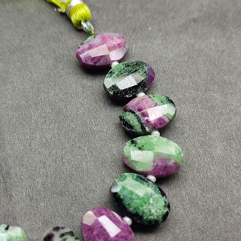Puede incluir: Un collar de cuentas ovaladas facetadas en tonos magenta intenso y verde. Las cuentas est&aacute;n ensartadas con un cord&oacute;n verde claro y amarillo. Las cuentas miden aproximadamente 2,5 cm de largo.