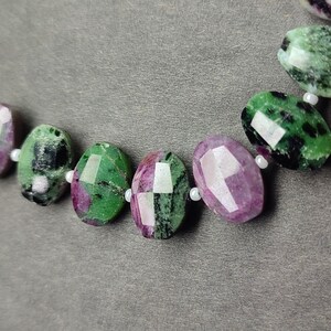 Puede incluir: Primer plano de un collar con cuentas ovaladas facetadas. Las cuentas alternan entre verde con inclusiones negras y rosa-p&uacute;rpura. Peque&ntilde;as cuentas blancas separan las piedras m&aacute;s grandes. El fondo es gris liso.