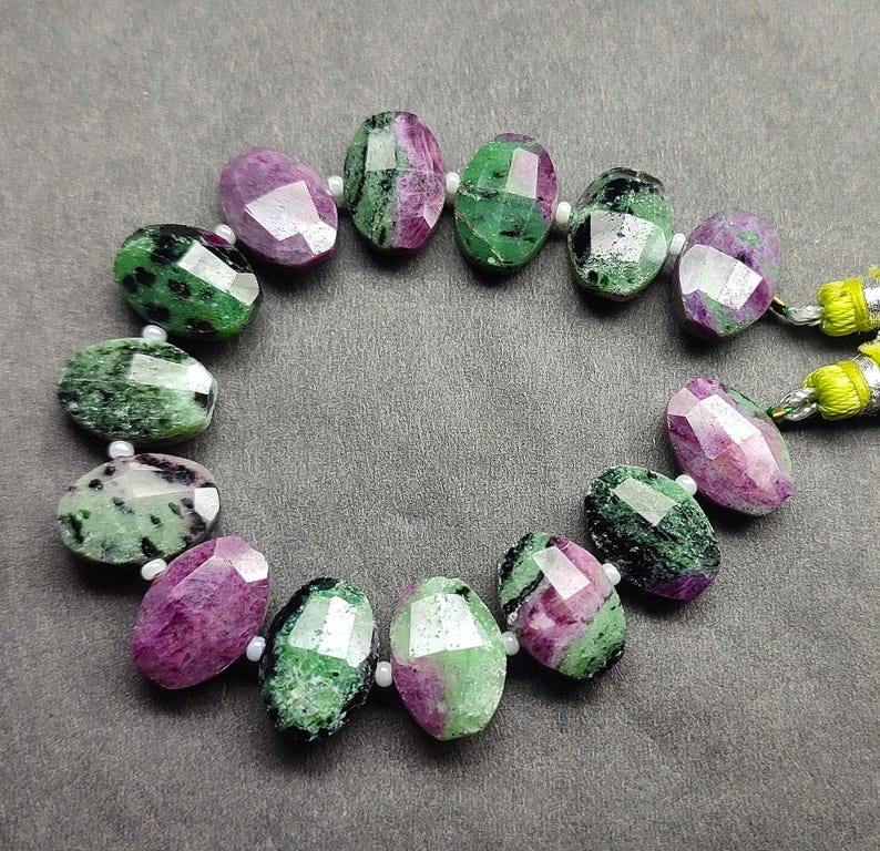 Puede incluir: Un collar de cuentas de piedras preciosas facetadas. Las cuentas de forma ovalada son una mezcla de verde y morado. Las cuentas est&aacute;n ensartadas con peque&ntilde;as cuentas blancas y un cord&oacute;n amarillo y verde.