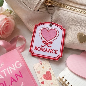 May include: A white rectangular embroidered patch with a scalloped edge and a red border. The patch features a pink heart graphic with a red bow underneath and the word ROMANCE in red capital letters. It is attached to a metal keychain ring.