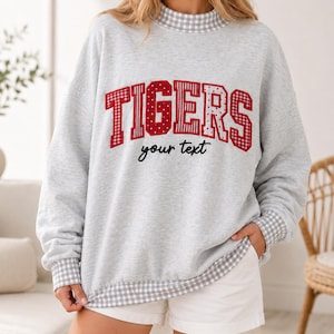 May include: A light gray long-sleeve sweatshirt featuring a mock neck collar, cuffs, and hem trimmed in a white and gray gingham pattern. The front displays the word TIGERS in large red letters with varied patterns and the text your text below.
