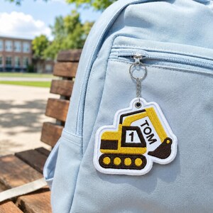 May include: A light blue backpack featuring a hanging embroidered keychain shaped like a yellow and black excavator. The excavator design includes the name TOM and the number 1 stitched in white thread on the side of the construction vehicle.