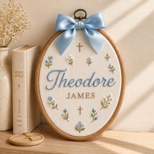 May include: An oval embroidery hoop featuring white fabric with the name Theodore James stitched in blue and brown thread. The design includes small blue and white floral sprigs, two small brown crosses, and a light blue satin ribbon bow on top.