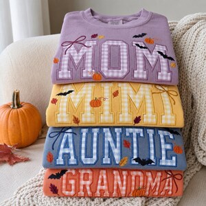 May include: A stack of four crewneck sweatshirts in purple, yellow, blue, and orange. Each features large block lettering reading MOM, MIMI, AUNTIE, and GRANDMA in a gingham pattern, decorated with embroidered pumpkins, autumn leaves, and black bats.