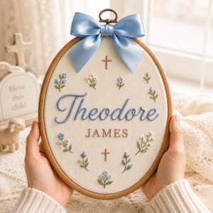 May include: An oval wooden embroidery hoop containing cream fabric with the name Theodore James embroidered in blue and brown thread. The design features small blue and white floral sprigs, two small brown crosses, and a large light blue satin bow.
