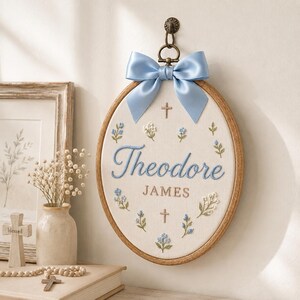 May include: An oval wooden embroidery hoop featuring the name Theodore James stitched in blue and brown thread. The fabric is decorated with embroidered blue flowers, green leaves, and two small brown crosses. A light blue satin bow hangs from the top.