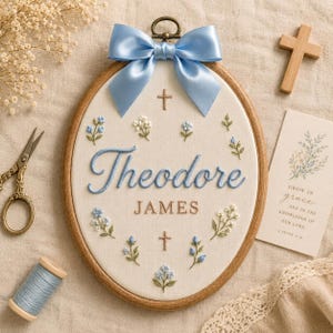 May include: An oval wooden embroidery hoop containing white fabric with the embroidered name Theodore James in blue and brown thread. The design features small blue and white floral sprigs, two small crosses, and a large light blue satin bow.