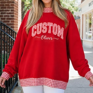 May include: A red long-sleeve sweatshirt featuring a mock neck collar, sleeve cuffs, and a bottom hem all trimmed in a red and white gingham pattern. The front displays the word CUSTOM in a block font and the name Oliver in a cursive script.