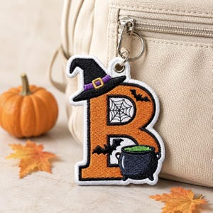 May include: An embroidered letter B keychain featuring an orange surface with a black witch hat on top, a white spider web pattern, two black bats, and a black cauldron filled with green liquid, attached to a silver metal keyring and clasp.