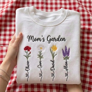 May include: White sweatshirt with the embroidered text "Mom's Garden" and four colorful flowers. Each flower has a name embroidered below it: Oliver, Sam, David, and Alisa. The sweatshirt is on a red and white checkered background.