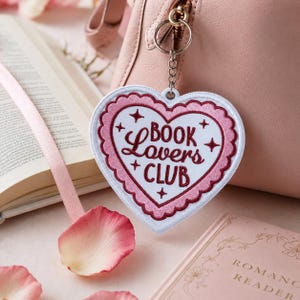 May include: A heart-shaped keychain featuring a white and pink scalloped border with the embroidered text BOOK LOVERS CLUB in dark red. The design includes small star accents and is attached to a silver metal ring and chain, hanging from a pink bag.