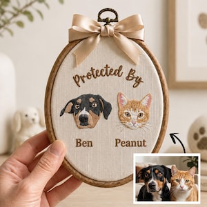 May include: An oval wooden embroidery hoop featuring a beige fabric background with the embroidered text Protected By, a black and brown dog portrait labeled Ben, and an orange tabby cat portrait labeled Peanut, topped with a beige satin bow.
