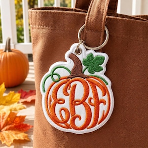 May include: A pumpkin-shaped keychain featuring orange embroidery with a white border and a brown stem. The center of the pumpkin displays the embroidered letters EBF in a cursive font. A green leaf and vine are attached to the top stem.