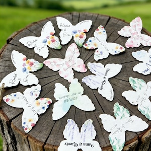 Puede incluir: Recuerdos de papel de semillas con forma de mariposa con diseños florales y nombres, dispuestos sobre un tocón de árbol. Cada mariposa tiene un patrón floral único y texto personalizado, ideal para bodas o eventos. Los nombres incluyen Rachel & Eric, Lucy & Frank y Anna & Randy.