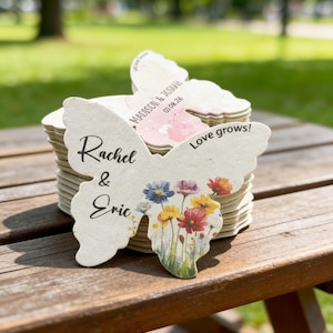 May include: A stack of butterfly-shaped paper cutouts with floral designs and names. One cutout features the names "Rachel & Eric" and a floral illustration. Another says "Madison & Joshau" with a date. The phrase "Love grows!" is also visible.