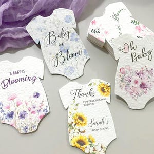 May include: A collection of onesie-shaped paper tags featuring floral designs and printed text including Baby in Bloom, A Baby is Blooming, Oh Baby, and Thanks for celebrating with us, Sarah's Baby Shower, with varied flower illustrations.