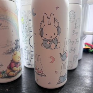 May include: A collection of white insulated water bottles, each decorated with cartoon illustrations. One bottle showcases a bunny with headphones, moons, and stars. Other bottles feature Winnie the Pooh and other characters.