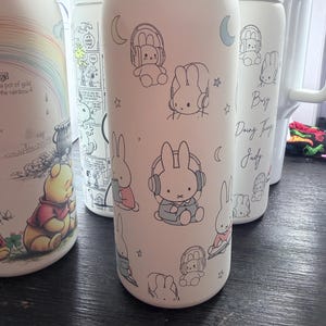 May include: White insulated water bottle featuring a pattern of cartoon bunnies wearing headphones. The bottle has a white lid and a silver base. Other bottles with cartoon characters are visible in the background.