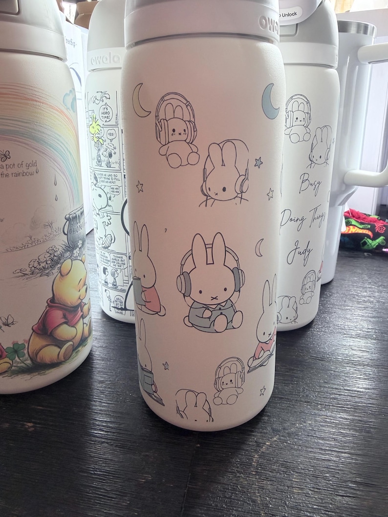 May include: White insulated water bottle featuring a pattern of cartoon bunnies wearing headphones. The bottle has a white lid and a silver base. Other bottles with cartoon characters are visible in the background.