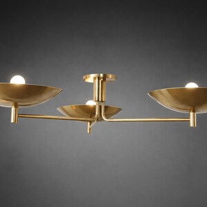 May include: A modern brass ceiling light fixture with three bowl-shaped shades. Each shade holds a small, round light bulb. The fixture has a central stem and horizontal arms extending to the shades, creating a balanced design.