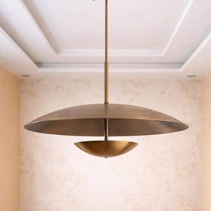 May include: A contemporary brass pendant light. It has a large, inverted disc-shaped shade above a smaller, bowl-shaped element. The light hangs from a slender rod attached to the ceiling, providing a warm, ambient light. The measurements are in centimeters.