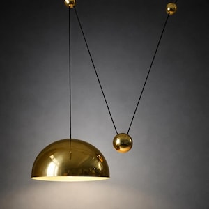 May include: A modern gold-colored pendant light with a half-dome shade and a smaller spherical accent. The light hangs from a black cord, creating a minimalist and elegant design. The fixture is suspended from the ceiling.