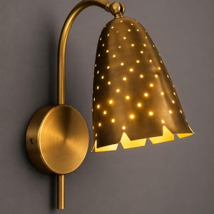 May include: A wall-mounted lamp with a brushed brass finish, featuring a bell-shaped shade with a perforated pattern that emits light through small circular holes. The fixture includes a circular wall plate and a curved arm with a hinge.