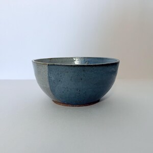 May include: A handmade ceramic bowl with a two-tone design. The bowl features a split color scheme, with a section of gray and a section of blue. The bowl has a rustic, textured appearance and a brown base.