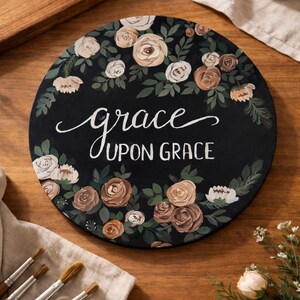 May include: Round, black decorative sign with the words "grace UPON GRACE" in white script. The sign is adorned with a floral wreath of cream, brown, and green roses. Paintbrushes and a small bouquet of flowers are nearby.