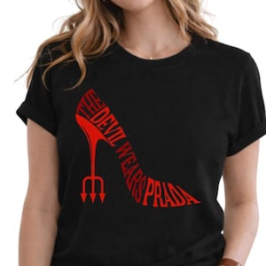 Pode incluir: Camiseta preta com um design de sapato de salto alto vermelho. A silhueta do sapato incorpora as palavras "THE DEVIL WEARS PRADA" em uma fonte estilizada, com um salto em forma de tridente.