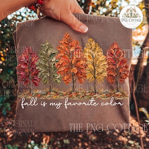 May include: A brown t-shirt featuring a graphic of five trees with autumn foliage in shades of red, green, orange, and yellow. Below the trees, the text "fall is my favorite color" is printed in a white, cursive, script-style font.