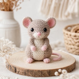 Op de afbeelding: Een handgemaakte gehaakte muis in grijstinten, beige en roze. De muis heeft grote zwarte ogen, roze oren, een roze neus en een gekrulde roze staart. Hij zit op een ronde houten schijf.