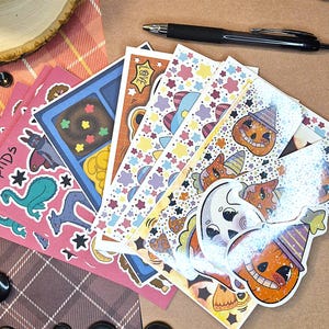 May include: A collection of rectangular sticker sheets featuring various colorful illustrations including a smiling ghost, jack-o'-lanterns in party hats, a teal dinosaur, and star patterns, arranged in a fan shape with a black pen resting on top.