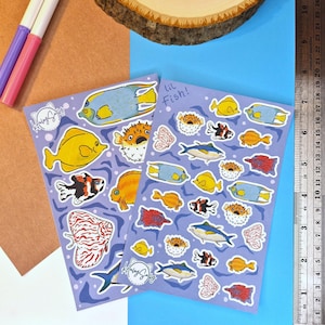 May include: Two rectangular sticker sheets with a light purple background featuring various colorful fish illustrations. The sheets display diverse species including pufferfish, angelfish, and tuna. One sheet includes the handwritten text lil Fish!.