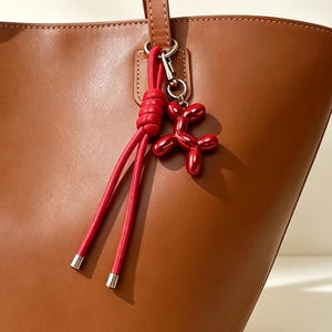 May include: A smooth brown tote bag with two shoulder straps, featuring a decorative red knotted cord charm and a metallic red balloon dog figurine attached to a silver-toned metal ring near the base of one strap.