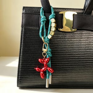 May include: A black textured handbag with a gold-tone metal clasp and a black fabric bow. Attached to the handle is a teal rope charm featuring gold-tone letter beads spelling SURF and a glossy red balloon dog pendant with a gold-tone clip.