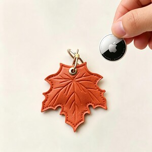 May include: A burnt orange leather keychain shaped like a maple leaf with embossed vein details and a gold-toned metal ring. The keychain is positioned next to a circular white and black Apple AirTag for scale, showing the relative size of the item.