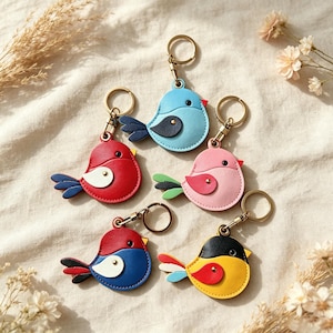 Könnte beinhalten: Fünf vogelformige Schlüsselanhänger aus farbigem Leder, jeweils mit einem Metallring versehen. Die Vögel sind in verschiedenen Farbkombinationen wie Blau, Rot, Rosa und Gelb gestaltet und haben kontrastierende Flügel, Schwänze und kleine schwarze Augen.