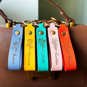 May include: Five leather keychains in light blue, yellow, turquoise, light pink, and orange, each featuring a gold metal snap, a gold ring, and a black engraved floral design with a name: James, Serena, Sak, Caitlin, and Victoria.