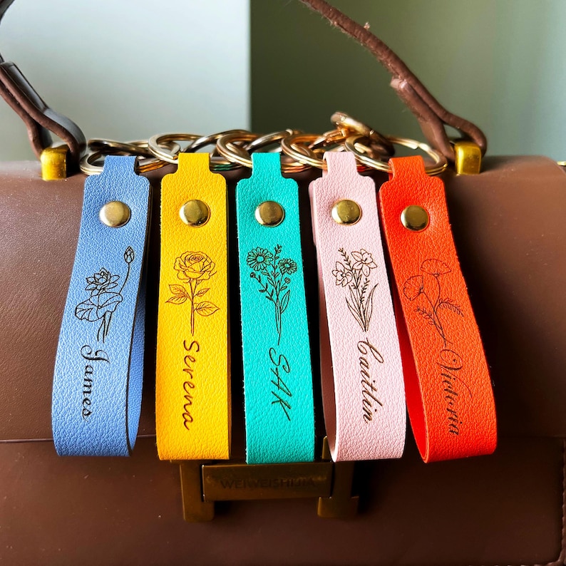 May include: Five leather keychains in light blue, yellow, turquoise, light pink, and orange, each featuring a gold metal snap, a gold ring, and a black engraved floral design with a name: James, Serena, Sak, Caitlin, and Victoria.