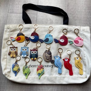 Könnte beinhalten: Eine Sammlung von fünfzehn vogel- und eulenförmigen Schlüsselanhängern aus lederähnlichem Material in verschiedenen Farben wie Gelb, Blau, Rot, Rosa und Grün. Jeder Schlüsselanhänger hat einen Metallring und liegt auf einer cremefarbenen Canvas-Tasche.