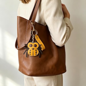Könnte beinhalten: Eine braune Ledertasche mit einem goldfarbenen Schlüsselanhänger aus Metall. Der Anhänger besteht aus einer gelb-dunkelbraunen Marienkäferfigur mit sechs Beinen und einem passenden gelben Lederband, das mit einer Rose und dem Text Serena geprägt ist.